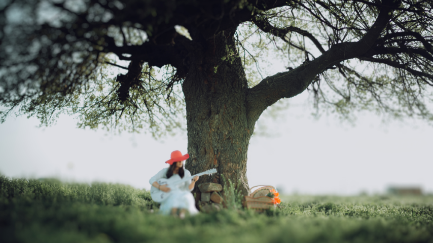 A woman in a peach colored hat and a white dress playing acoustic guitar under a tree